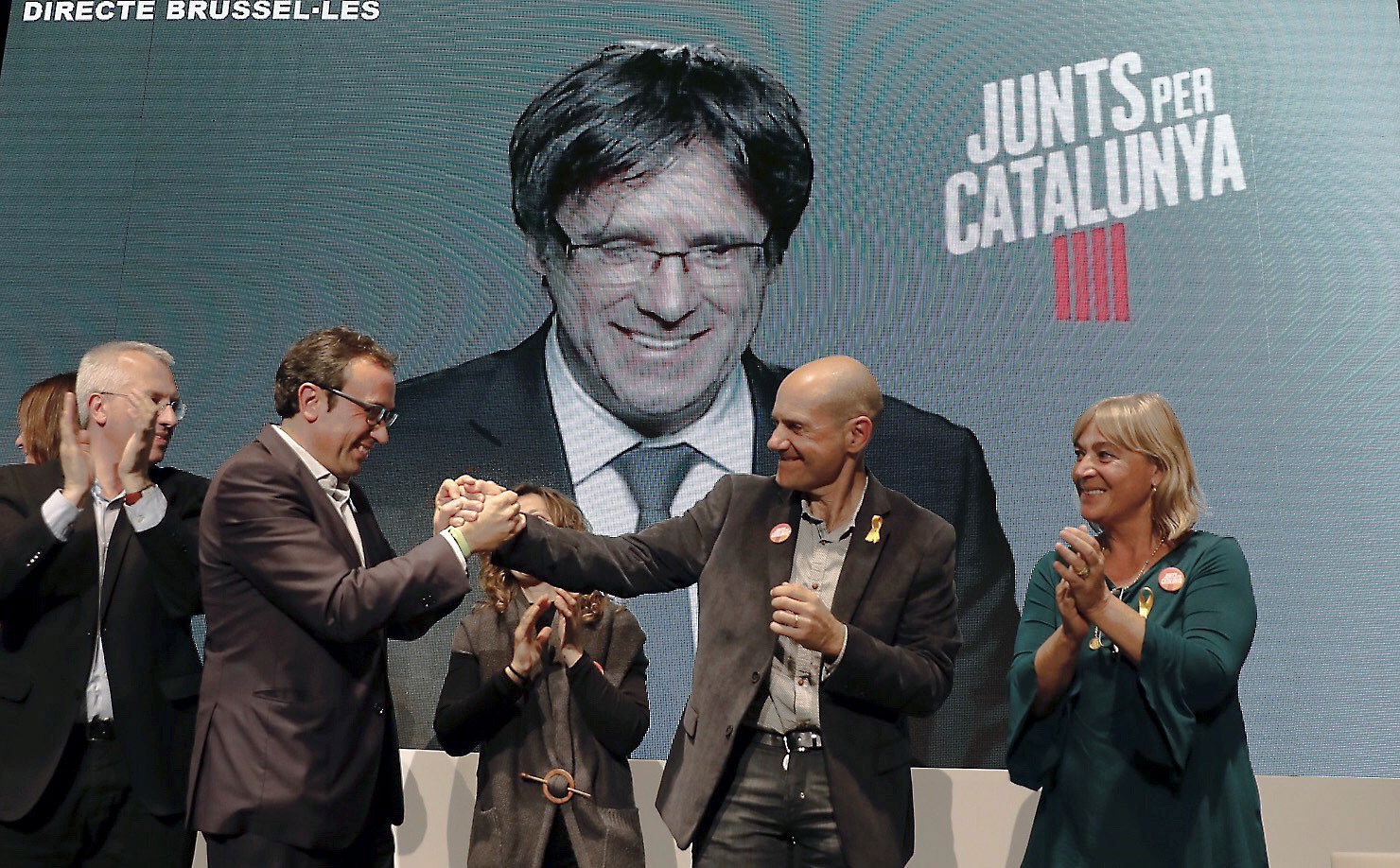 Junts per Catalunya's candidates Josep Rull (2ndL) and Xavier Quintillá (2ndR) shake hands as Catalonia's former president Carles Puigdemont (at the screen) speaks in a videoconference from Brussels during an electoral campaign event in Lleida, Catalonia, Spain, Dec. 18, 2017.